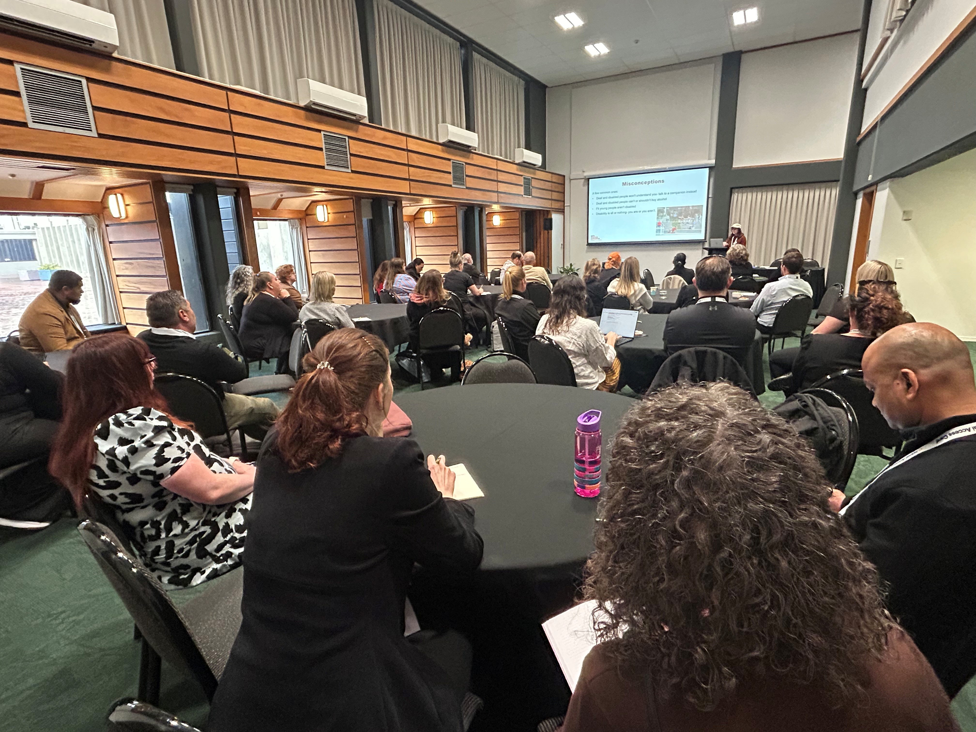 Attendees sit at round tables in a large hall while listening to Katie Querin deliver a Front of House Accessibility Training